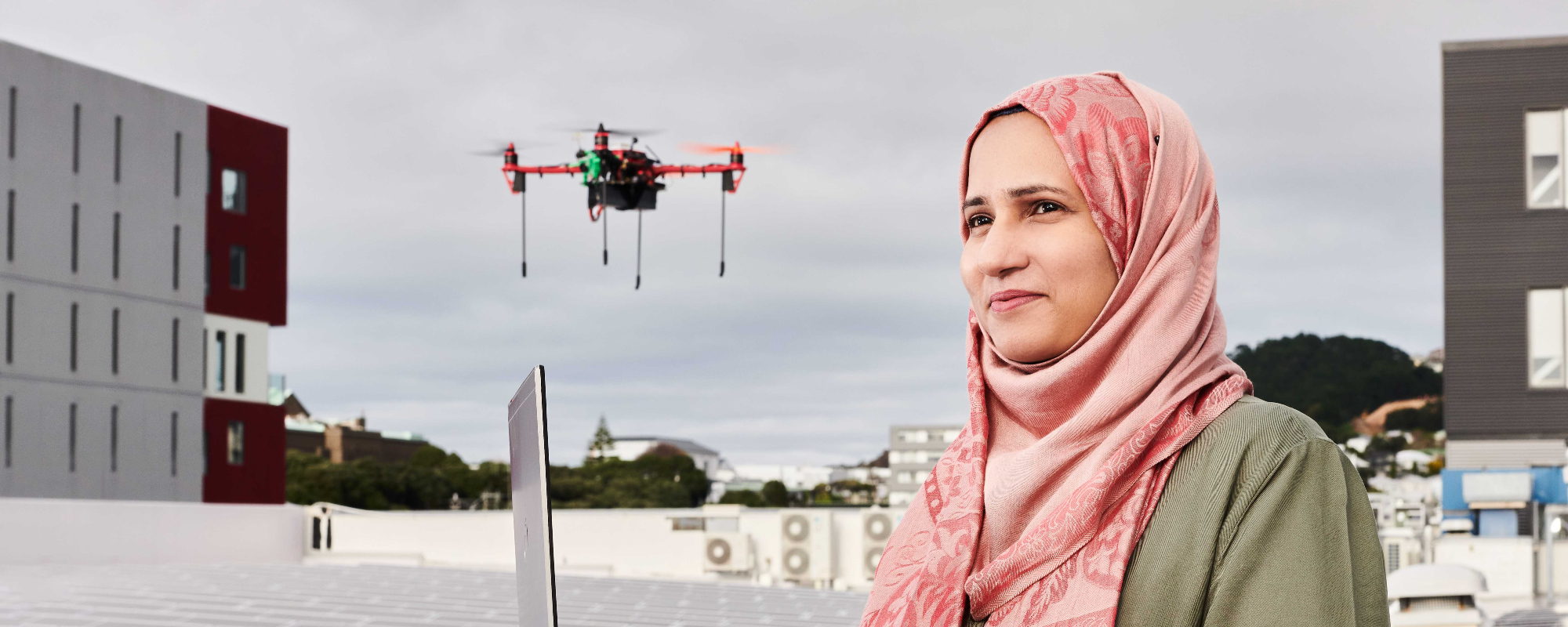 Master of Engineering graduate Rooman Khalid standing on Te Aro campus roof with a solar panel grid and a drone behind her, holding her laptop.