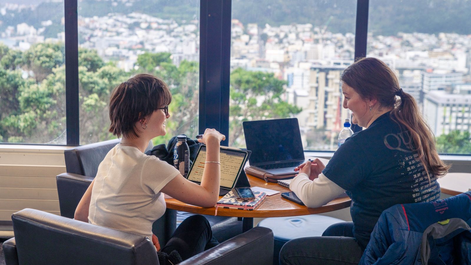 Two students sitting at a table in the Library with their laptops and notebooks, and a view of the city in front of them.