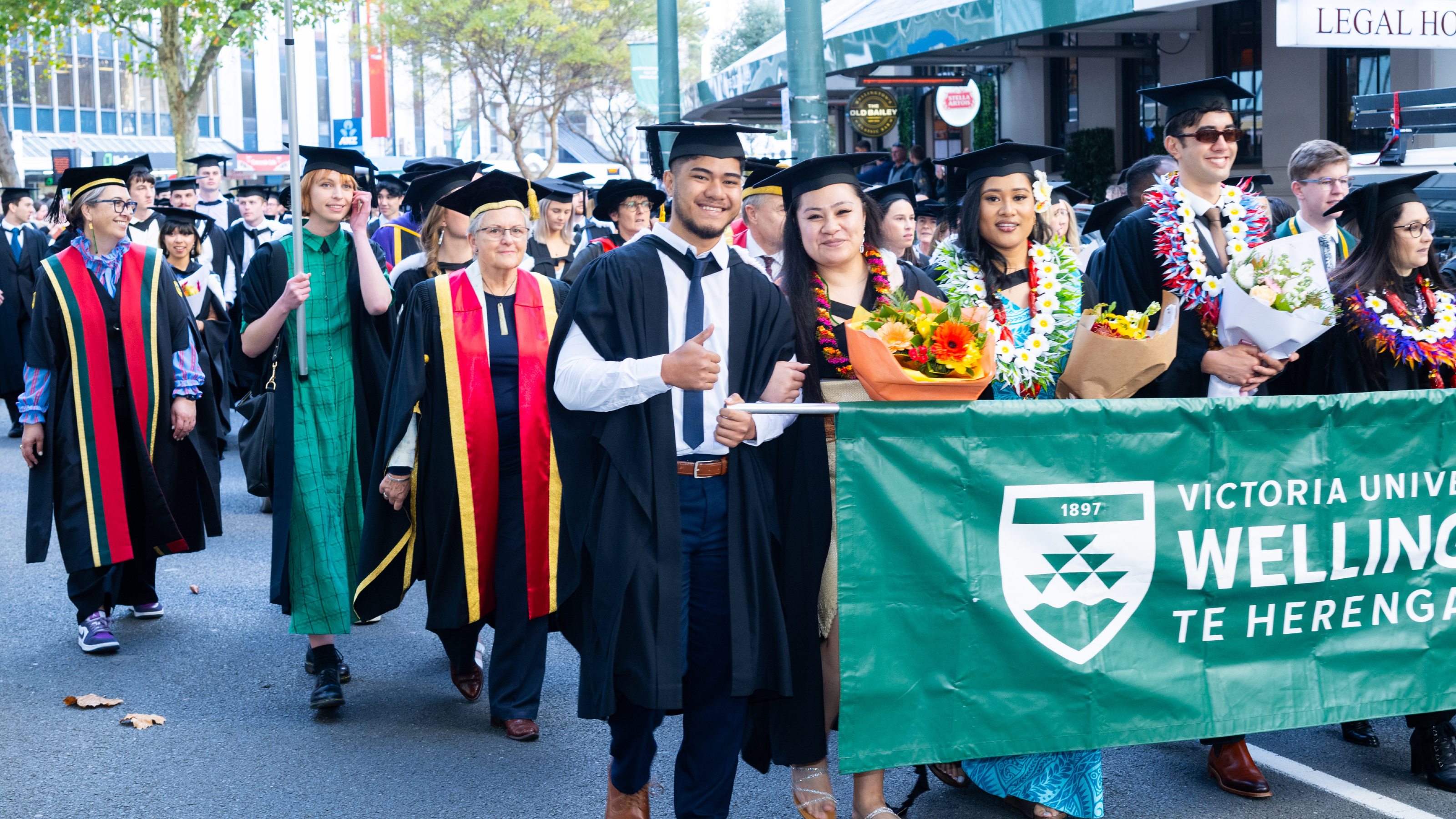 A picture of students in academic dress holding flags and a banner in a graduation parade in central Wellington. 