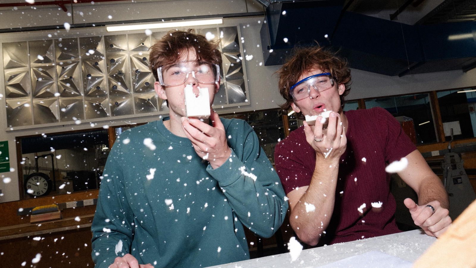 Two students wearing safety glasses blowing sanded wood dust toward the camera.