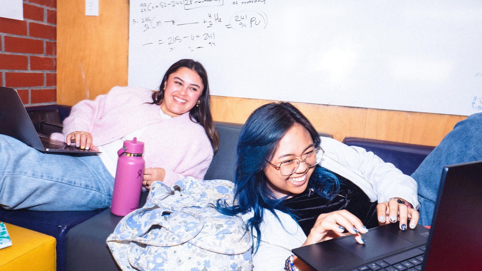 2 students lounging on a couch in front of a whiteboard with math formulas, with their laptops open