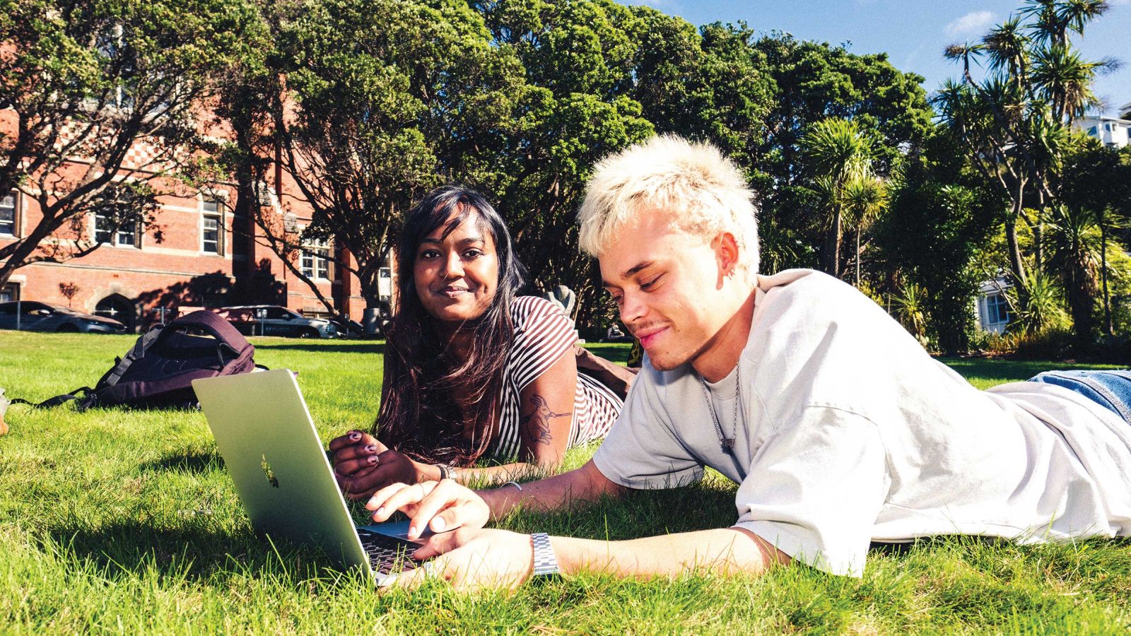 two students laying on grass outside university building studying on a laptop