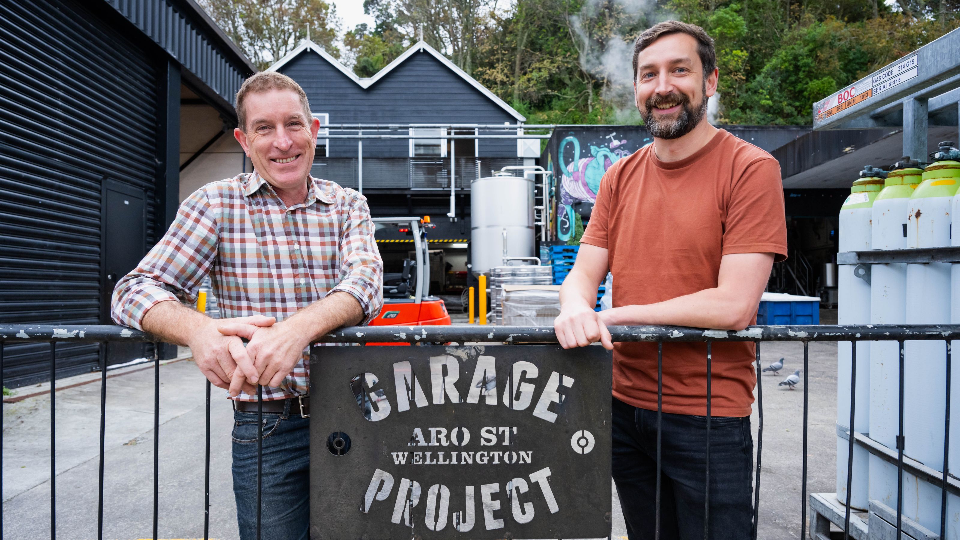 Simon Hinkley and Peter Bircham standing behind a metal railing with a sign reading ‘Garage Project Aro St Wellington,’ with industrial buildings and equipment in the background.