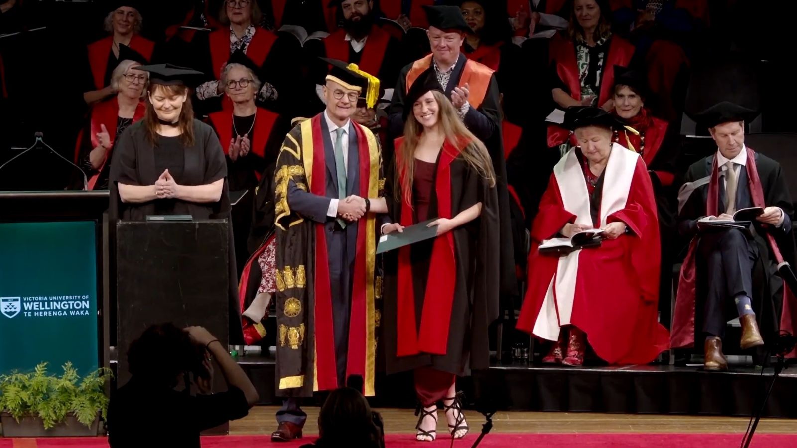 Graduate receiving a diploma and shaking hands with the Chancellor on stage during a ceremony.