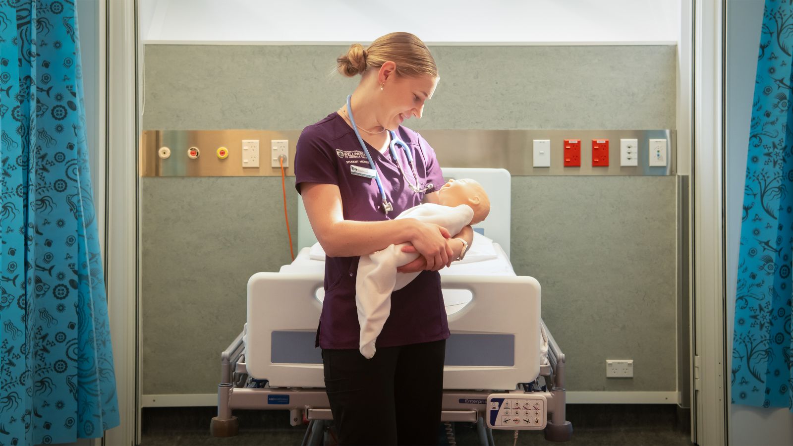 A bonde student in scrubs holds a baby doll in front of a hospital bed