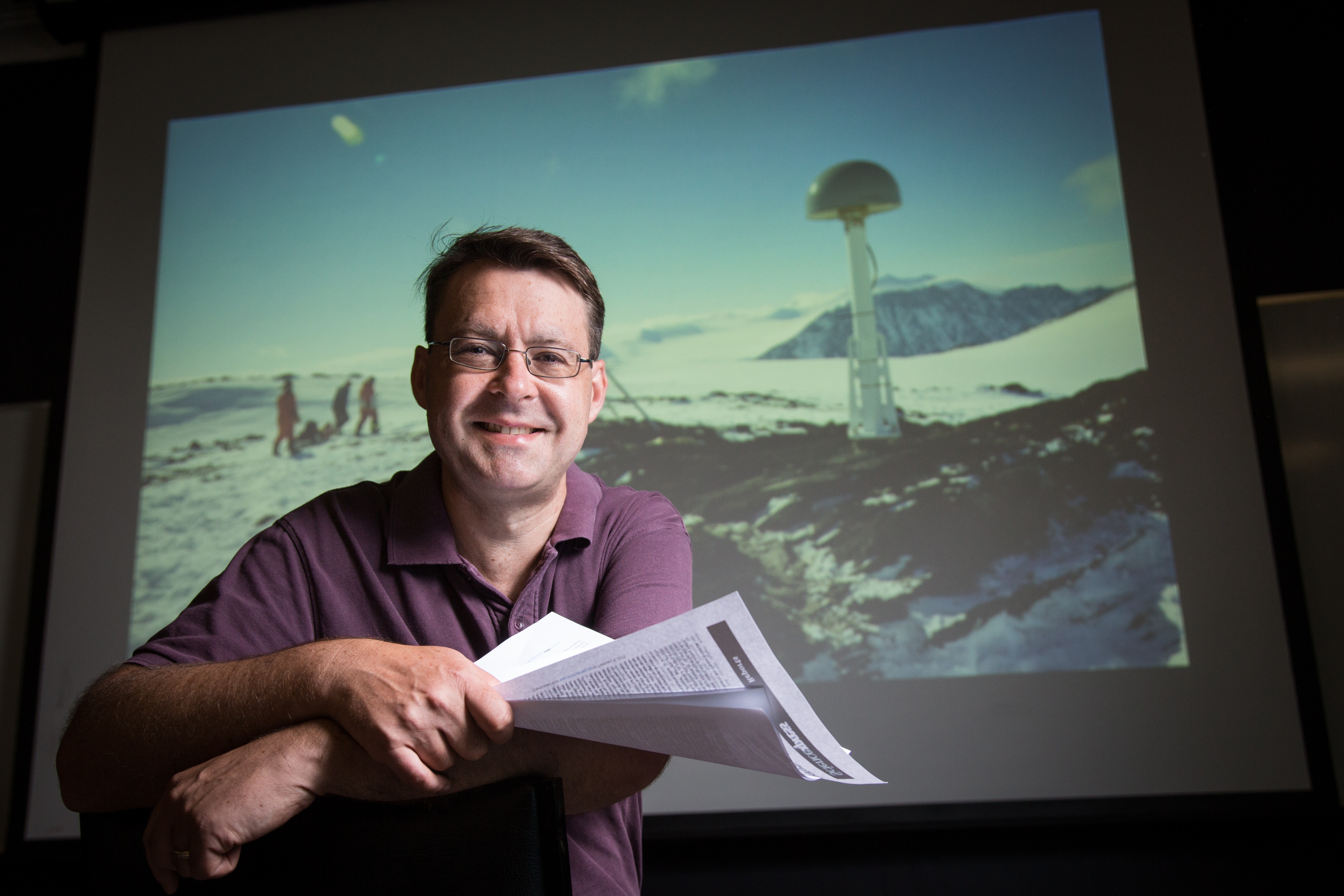Scientist Matt King stand in front of a projected image of Antarctica. He faces the camera with papers in his hand. 