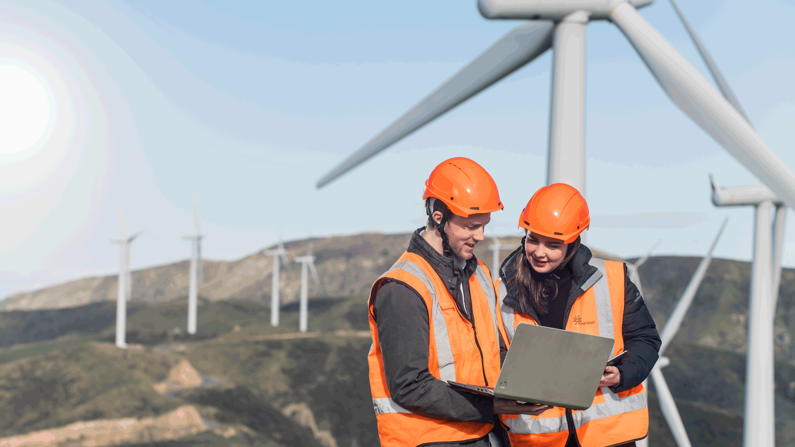 Two students wearing high visability orange vests and hard hats, stand outside on a hill with wind turbines in the background, and look at a laptop together.