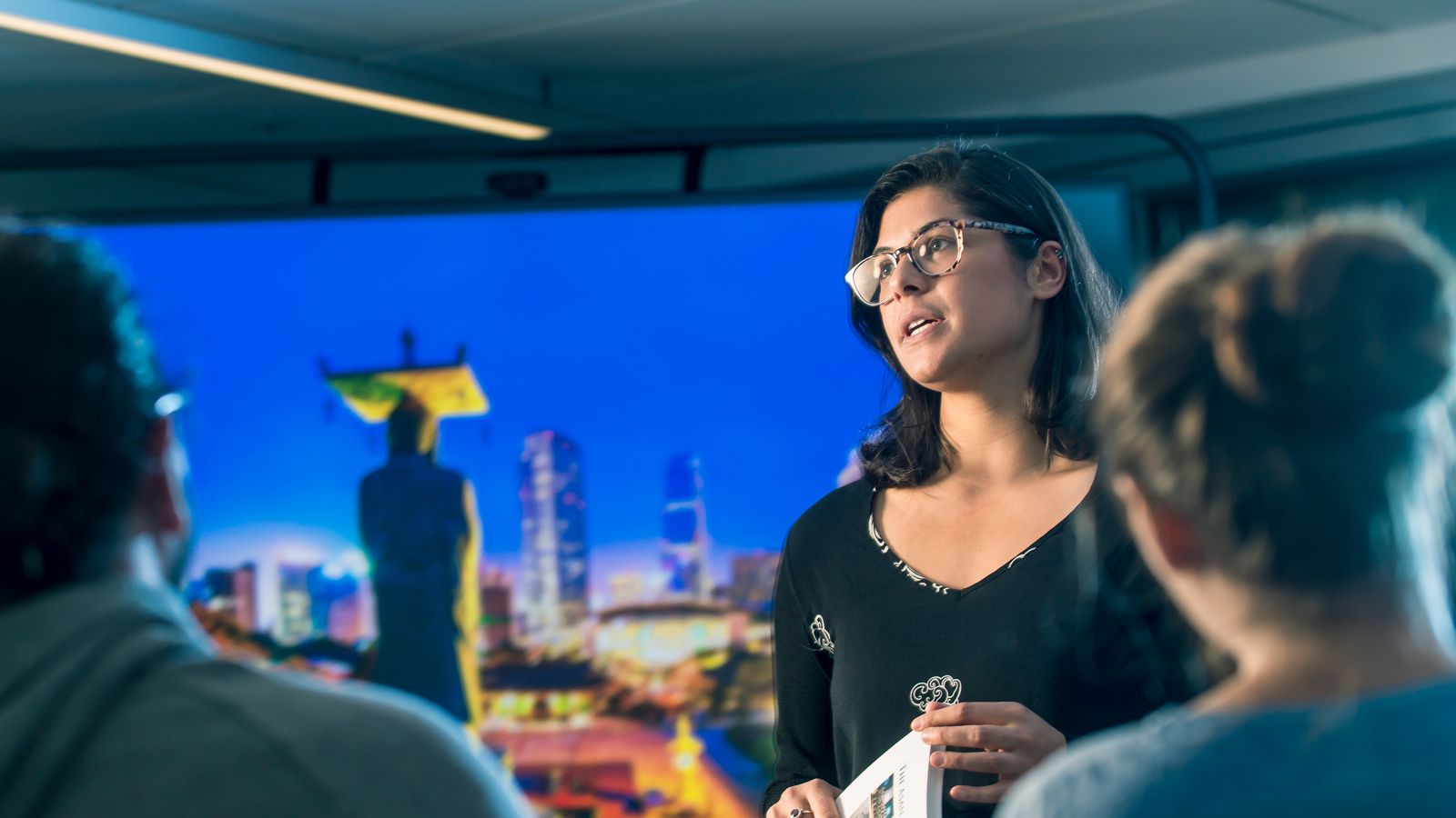 Woman addressing listeners, with city at night in background.