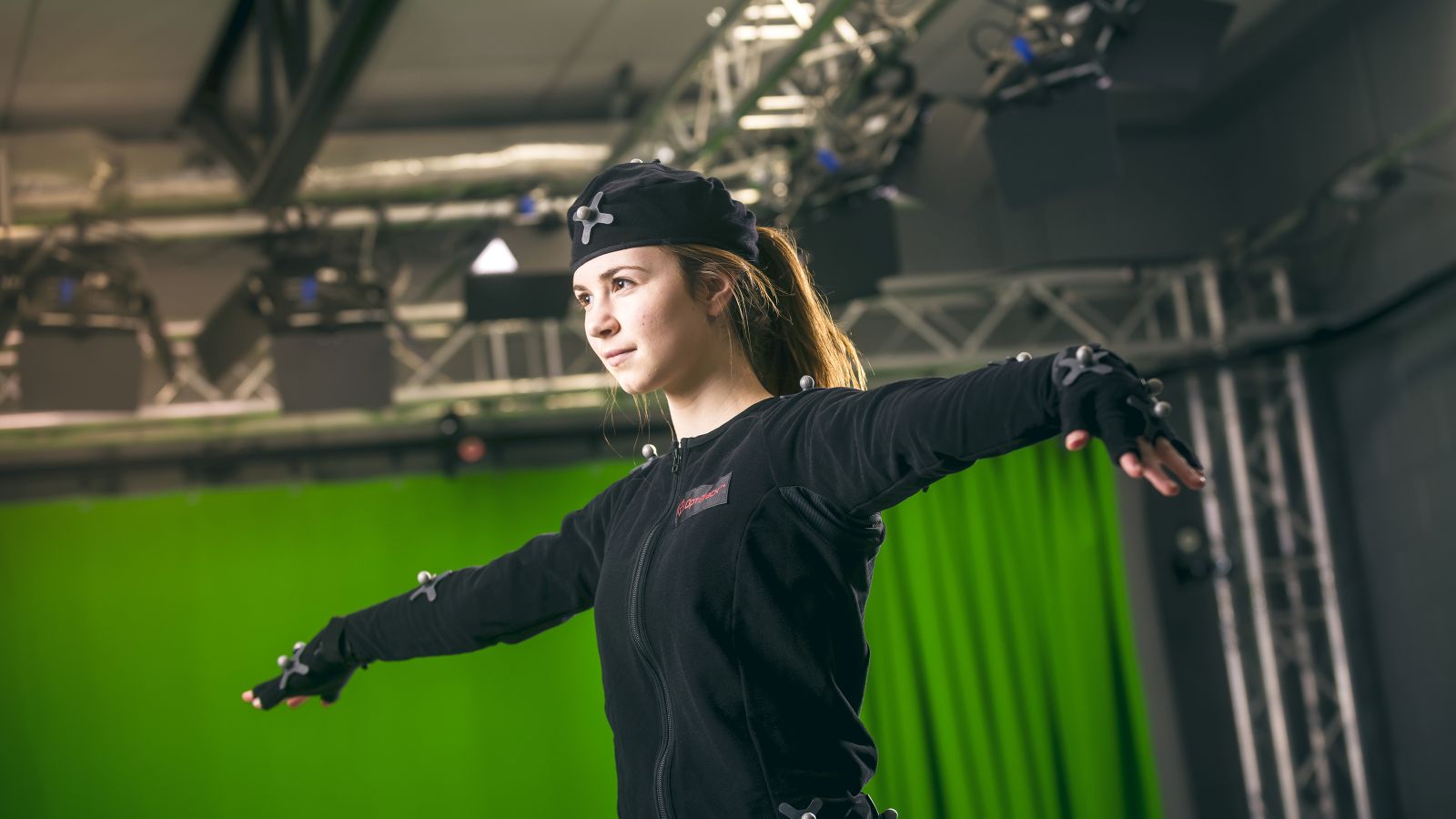 A female student in a motion capture student extends her arms in front of a green screen.