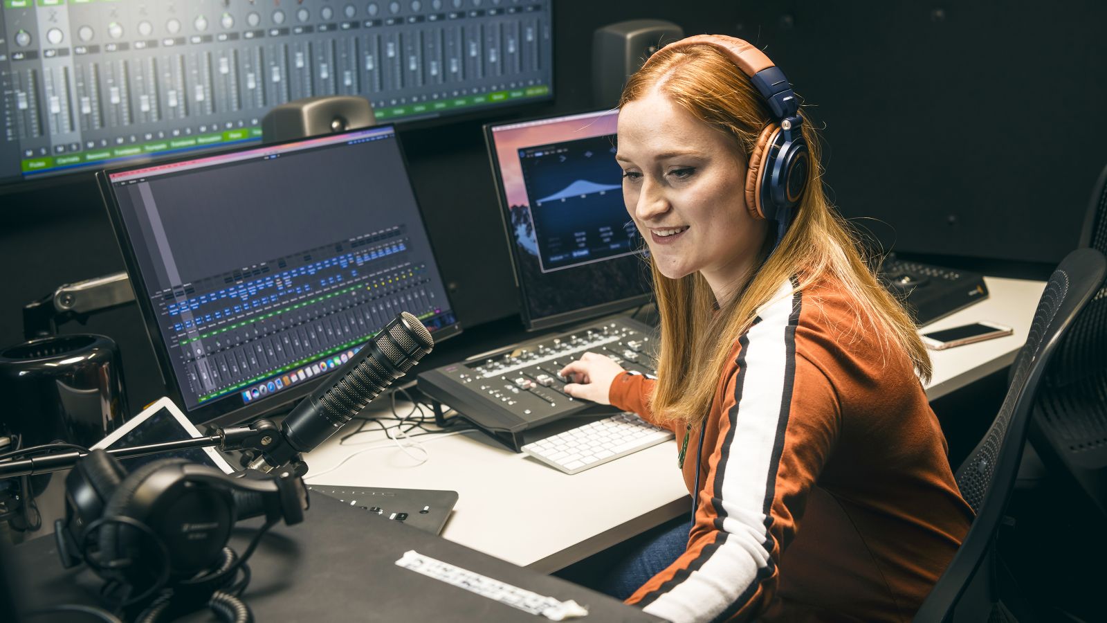 A female student with long red hair wears headphones while operating equipment in a recording studio.
