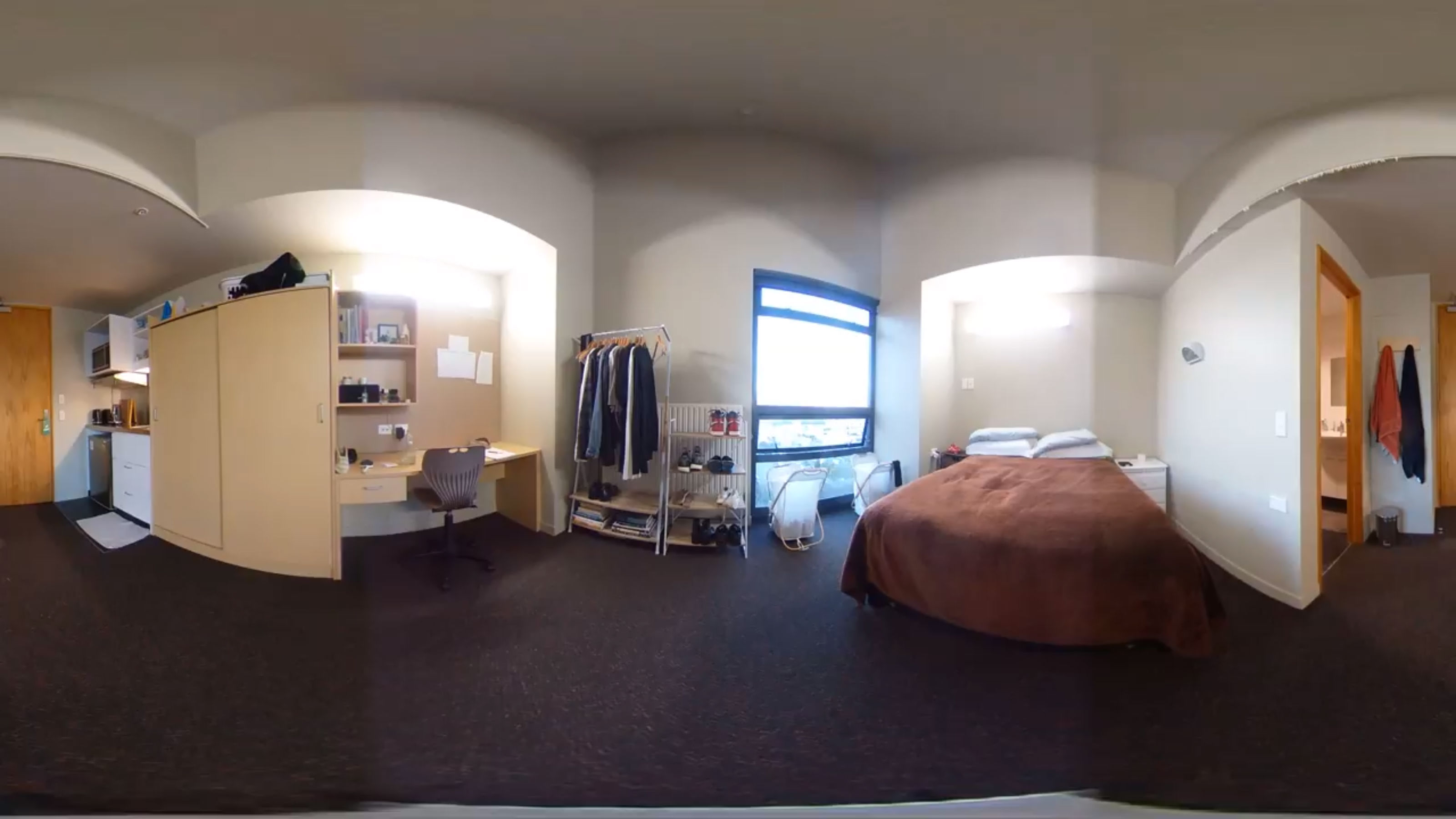 A panoramic view of an occupied bedroom with am ensuite, desk, double bed, and kitchenette.