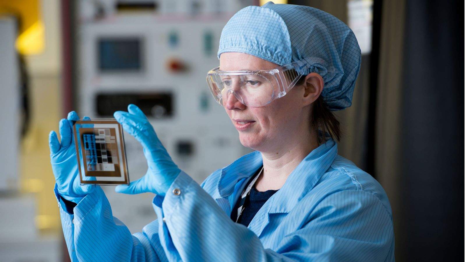 A bespectacled Dr Natalie Plank in a bouffant hair net and lab coat holds up for inspection, between latex-gloved hands, a transparent biosensor.