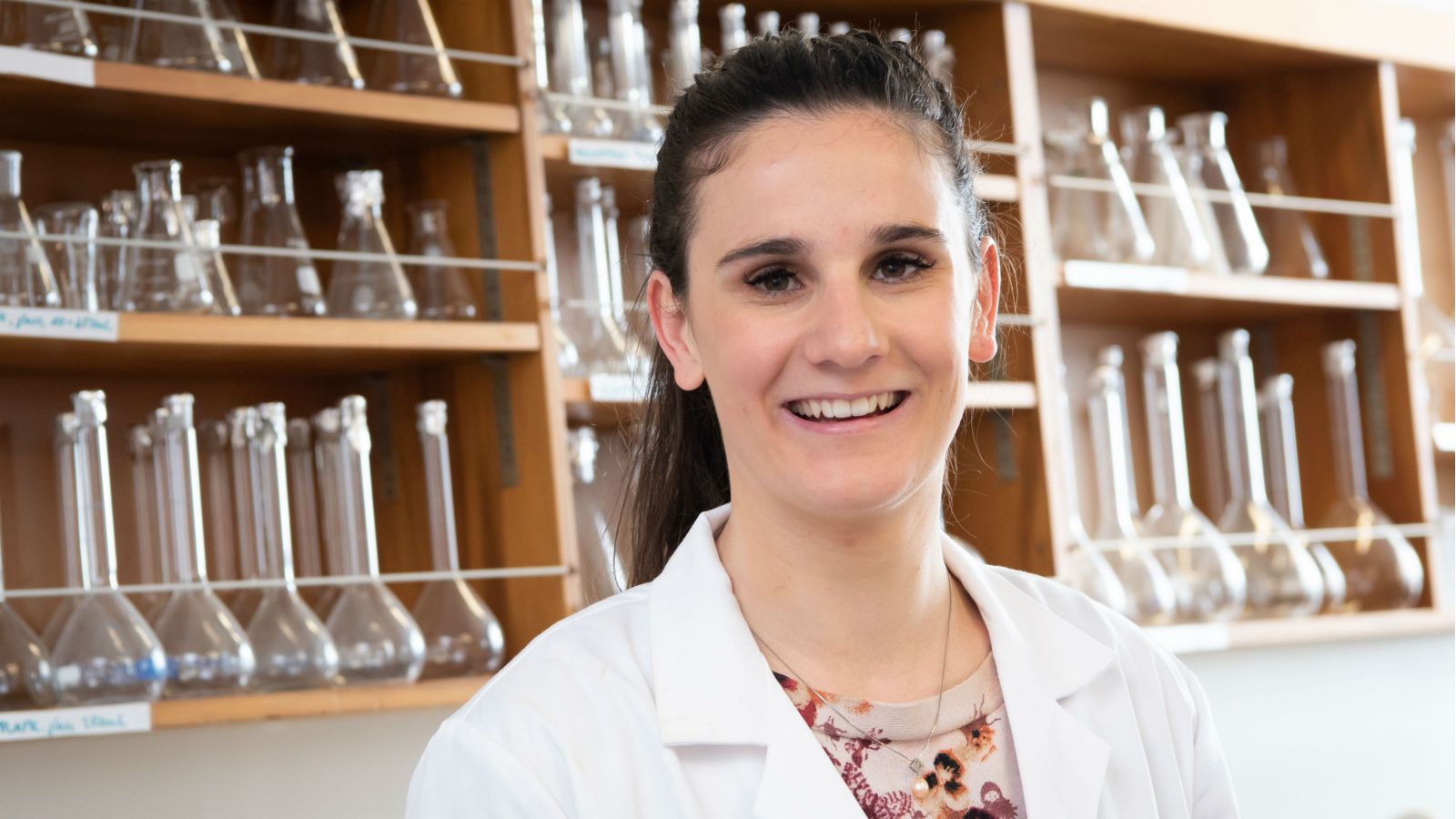 Francesca Mills, wearing a lab coat, stands in a lab in front of rows of glass beakers