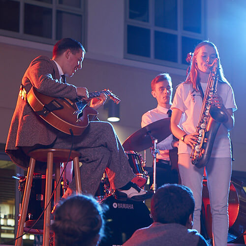 A jazz band, comprised of a guitarist, saxophonist, bassist, and drummer, play on a dim stage, illuminated by red and blue backlights. 