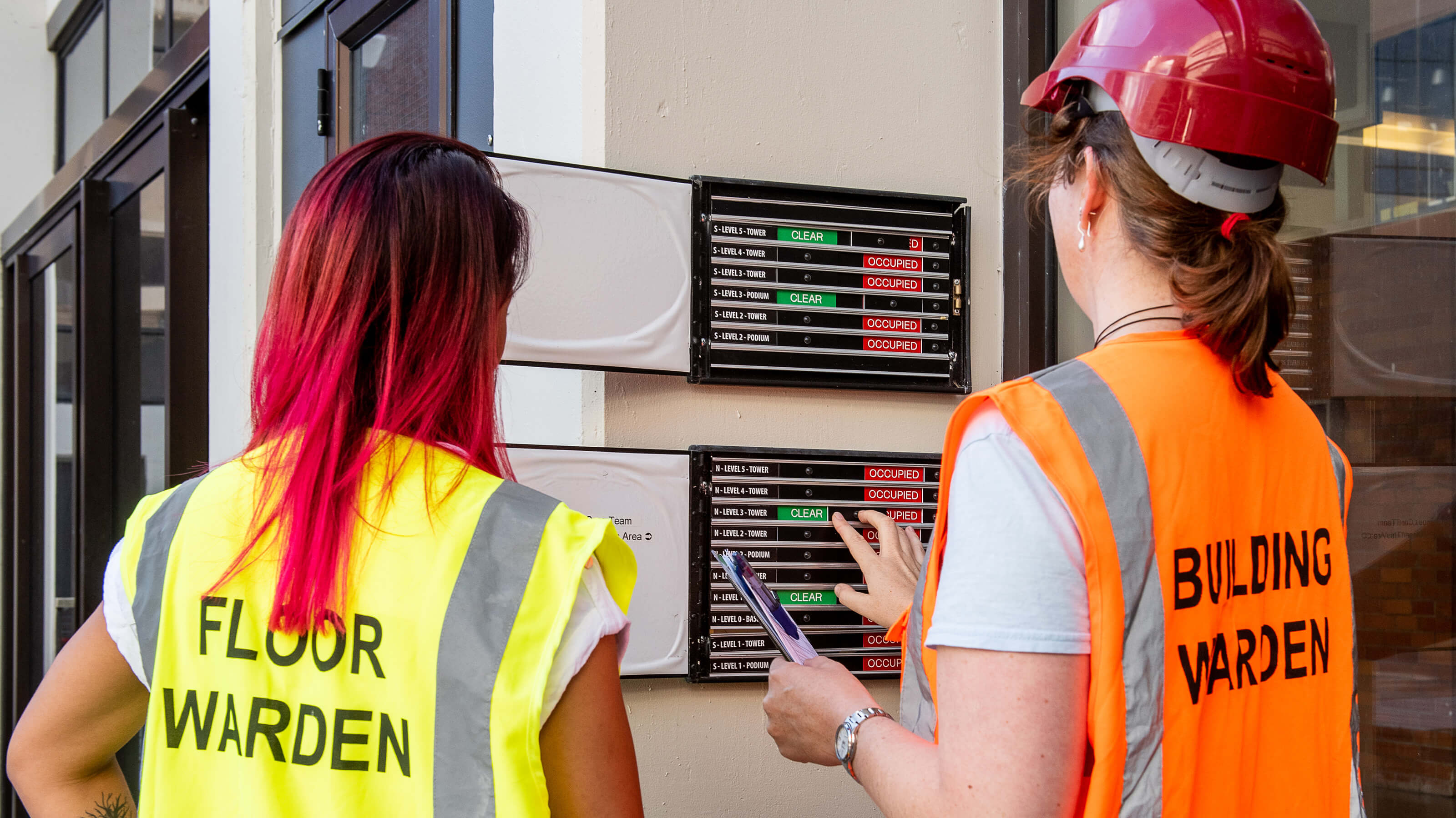 Two women looking at an evacuation board. One woman wears a yellow vest labelled floor warden and the other woman wears an orange vest labelled building warden.