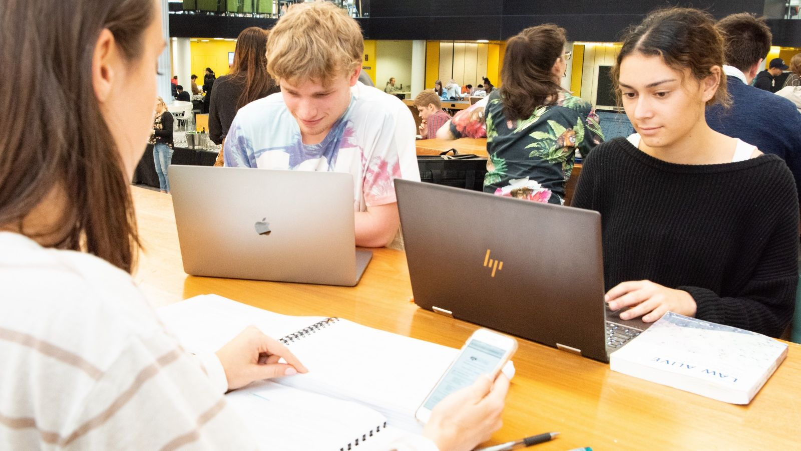 Students in the hub, two sitting at laptops, while one uses a smartphone.