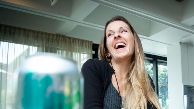 A woman laughs in while sitting at a table, a blurred silver soft drink can is in the foreground.