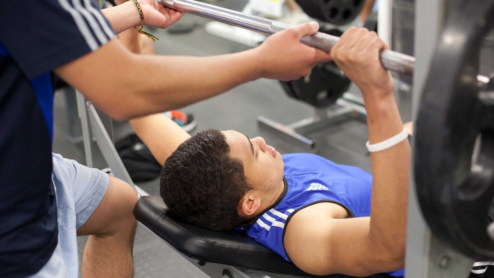A Māori man lifts weights with a spotter on a bench press.