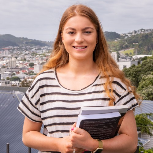 Smiling student, Olivia Watson, is standing on Kelburn campus with Wellington harbour in the background. 