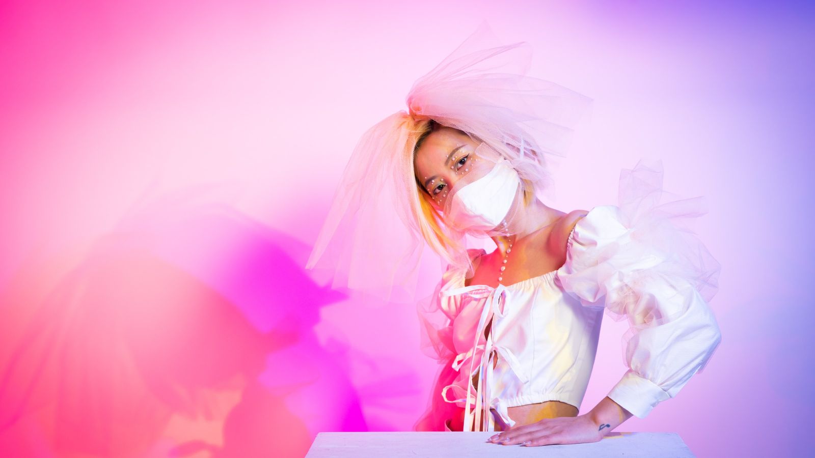 Model wearing tie front white shirt with dramatic organza headpiece in front of pink background