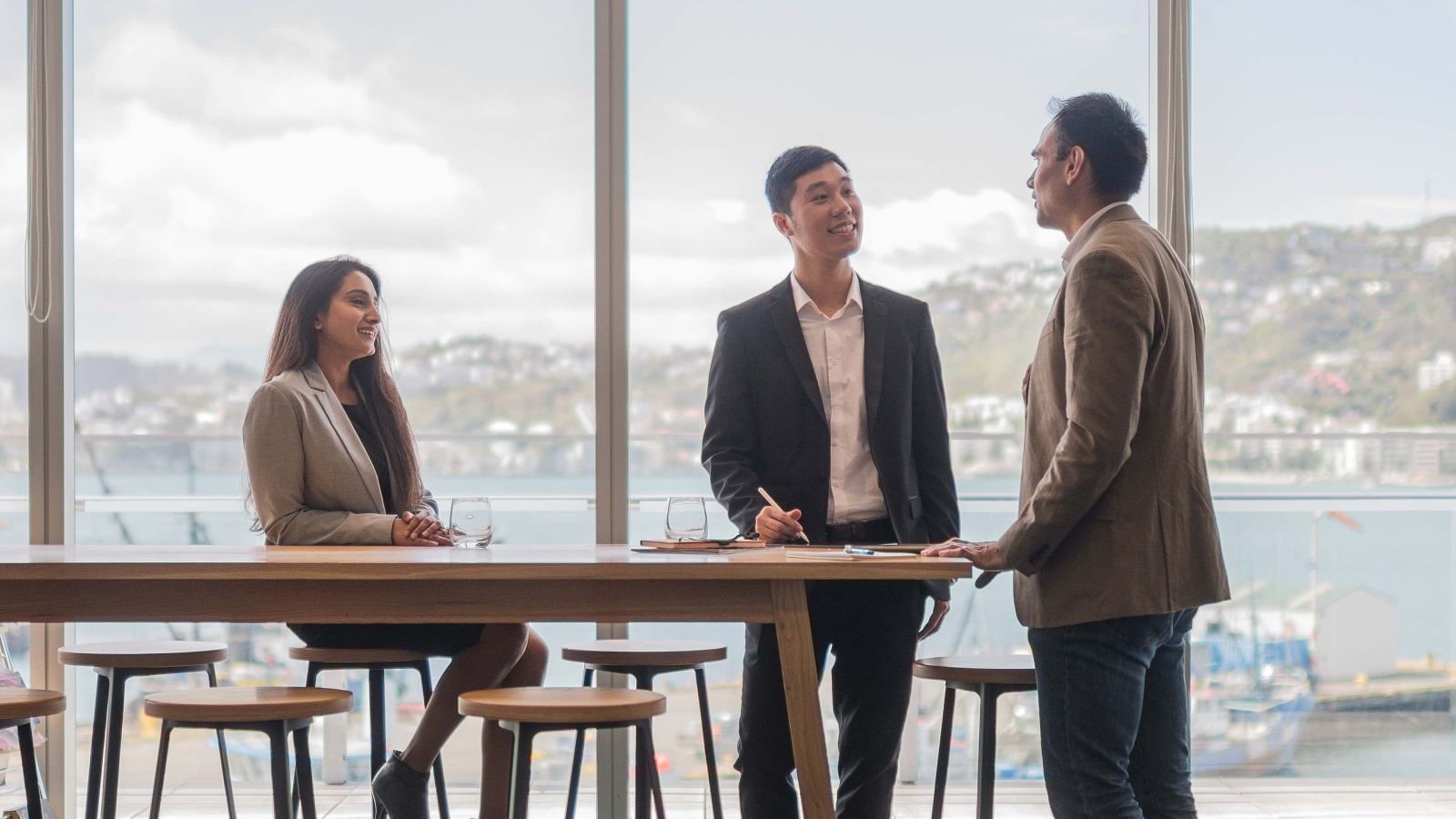 Three profesisonal programmes students meet in a modern office with Wellington Harbour in the background.