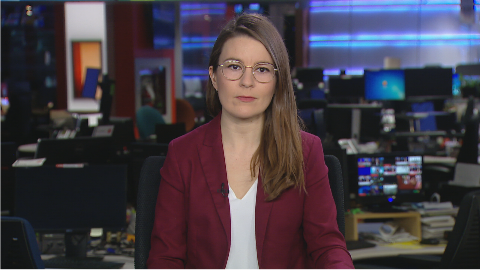 picture of girl with glasses, long hair, with white top and marron blazer sitting in front of a newsroom