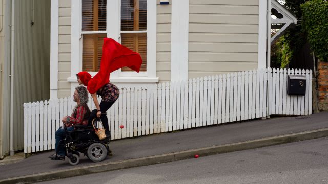 A man dressed as a wolf drives a motorised wheelchair down a steep street with a woman dressed like Little Red Riding Hood with a red cape and basket stands on the back.