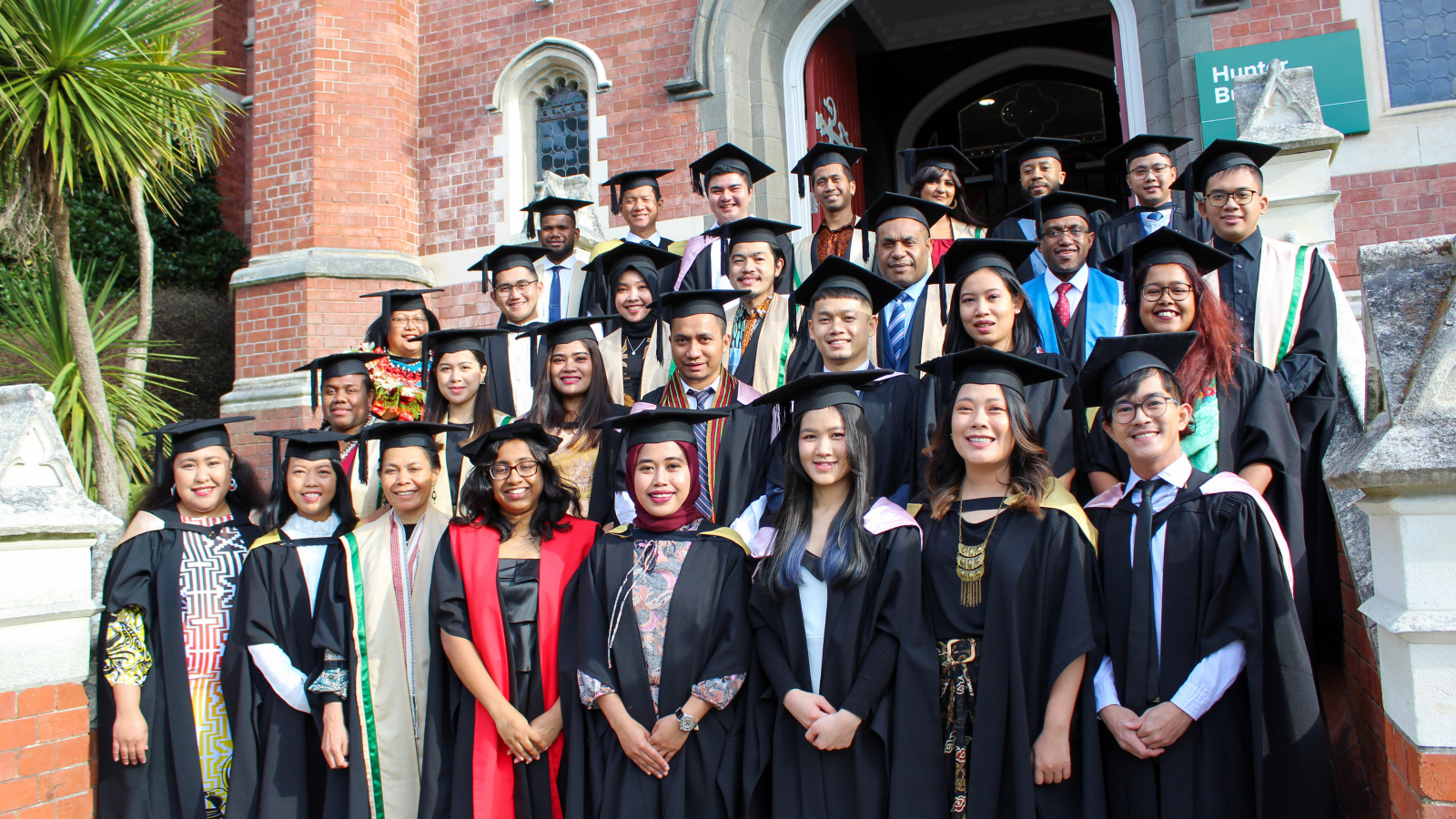 Group of 32 graduates on steps of gothic style university building