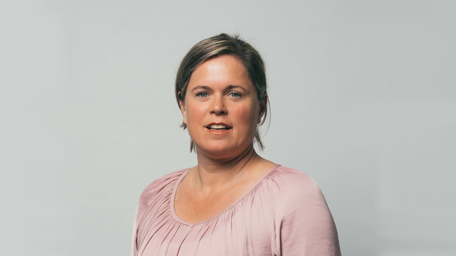 Bindi smiles at the camera wearing a pink top behind a grey background