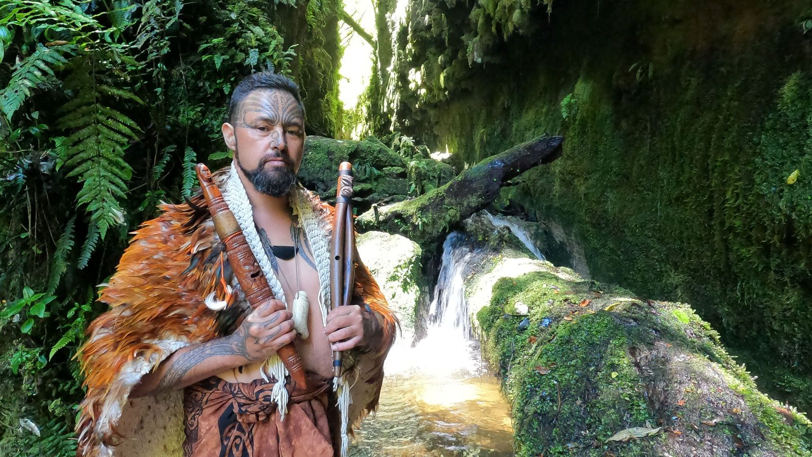 Man with māori cloak holding traditional taonga puoro instruments against natural background