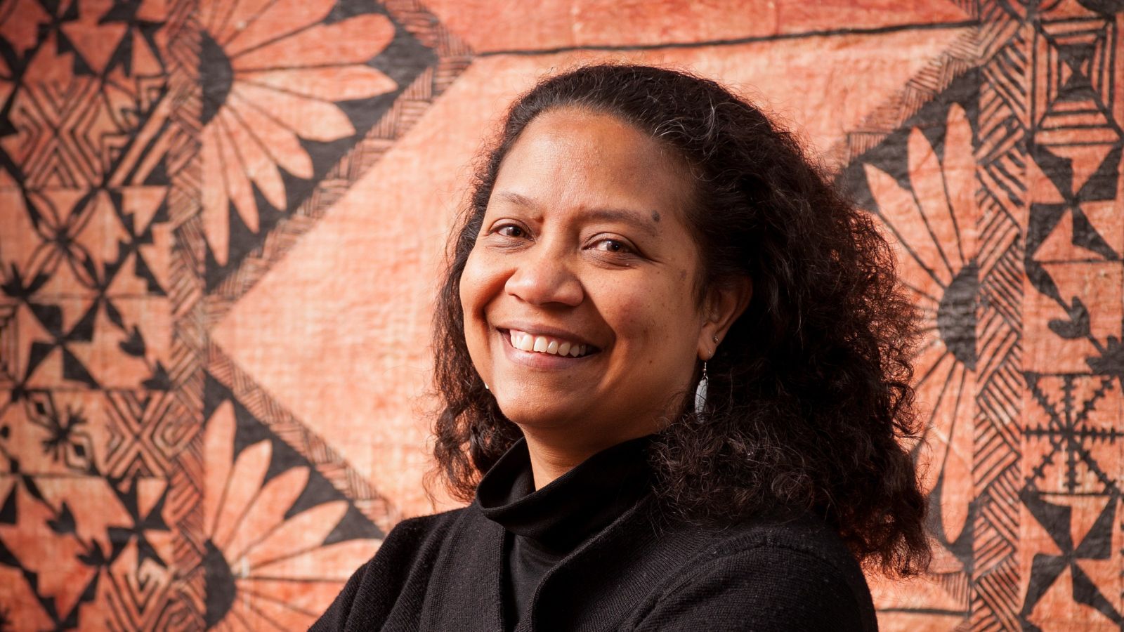 Teresia Teaiwa standing in front of a tapa cloth that is hanging on a wall.