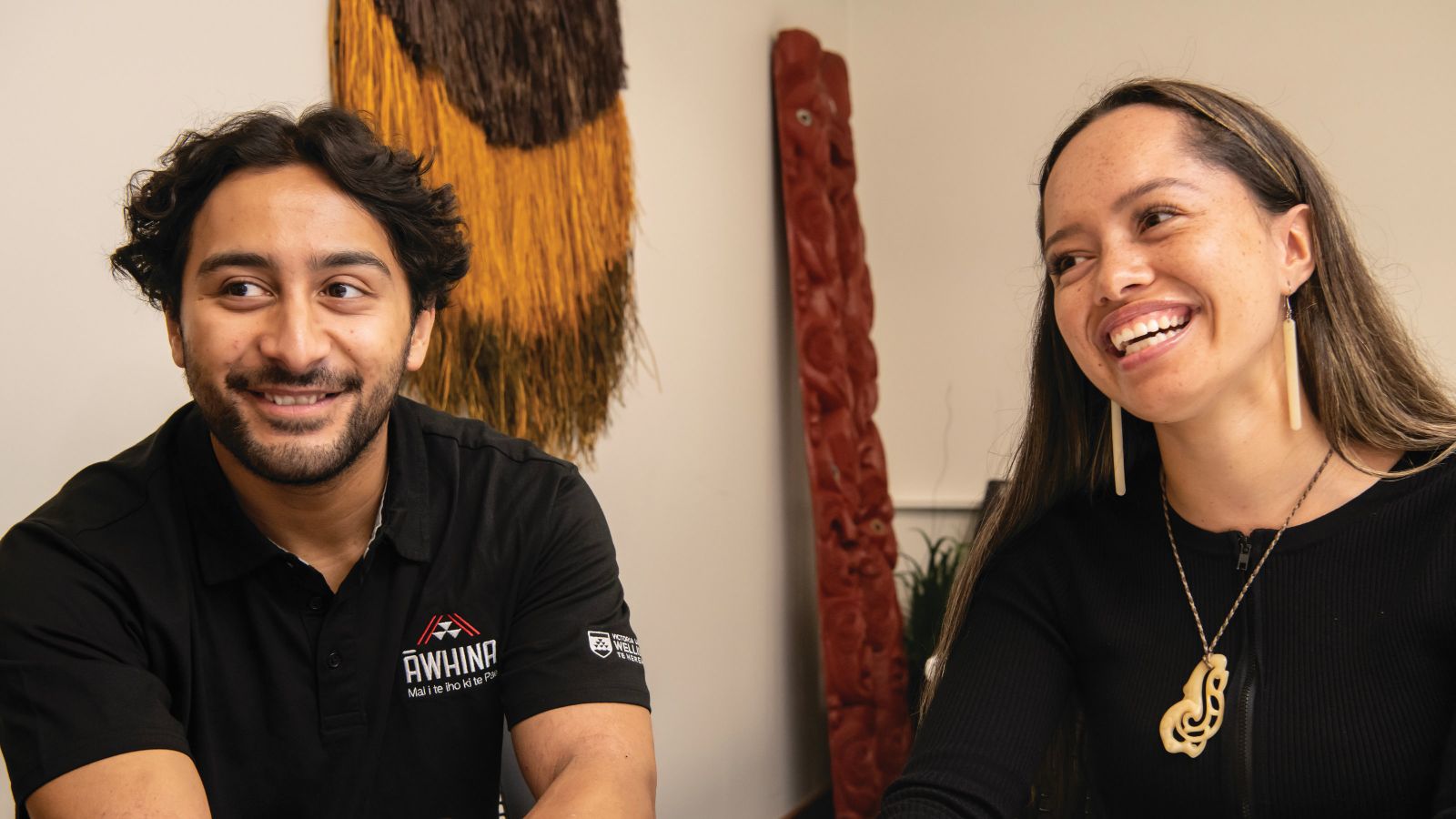 A young Māori man wearing a black shirt with an Awhina logo and a young Māori woman, both laughing. 