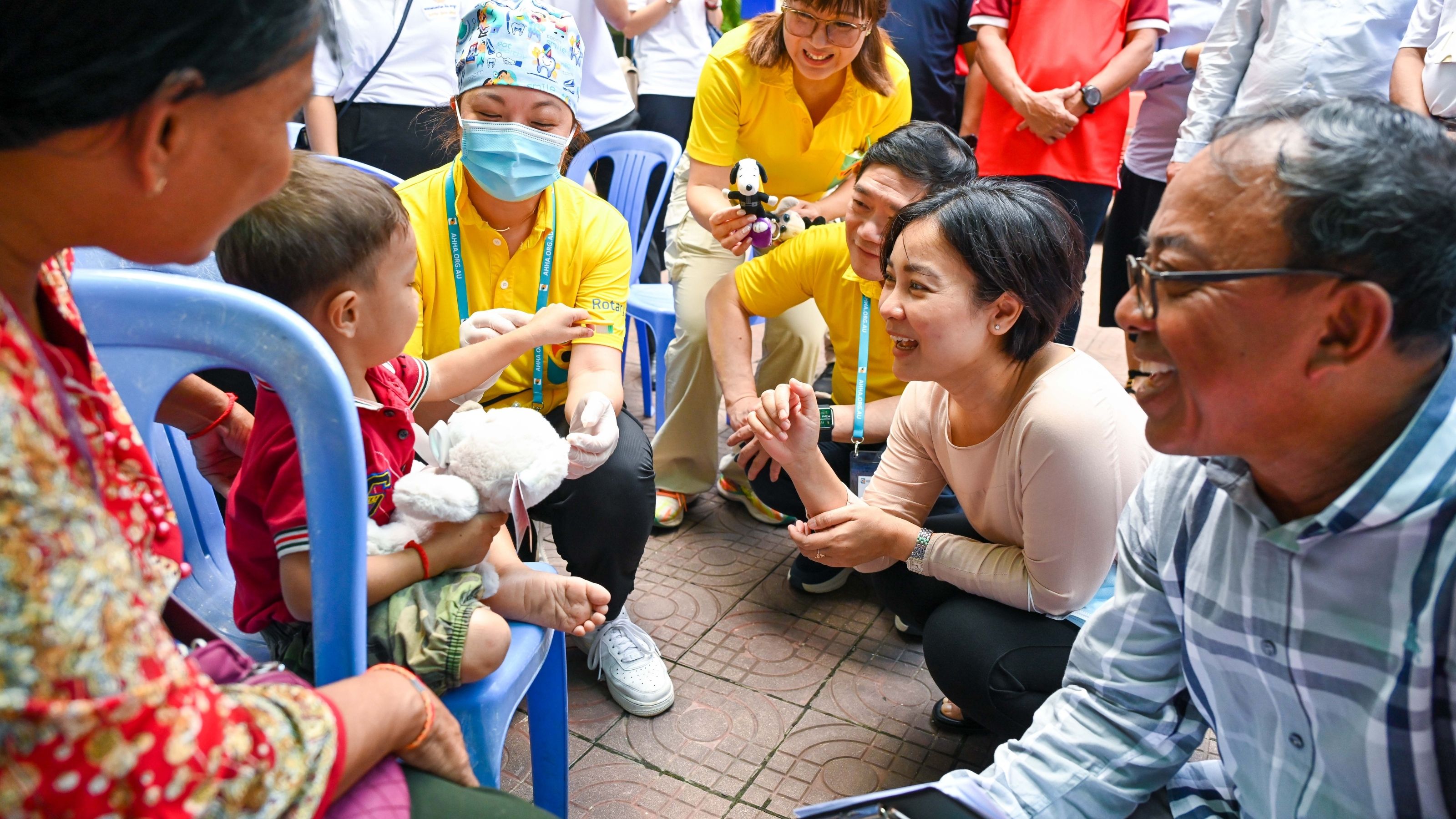 A group of people are gathered around a toddler on a chair getting immunised.