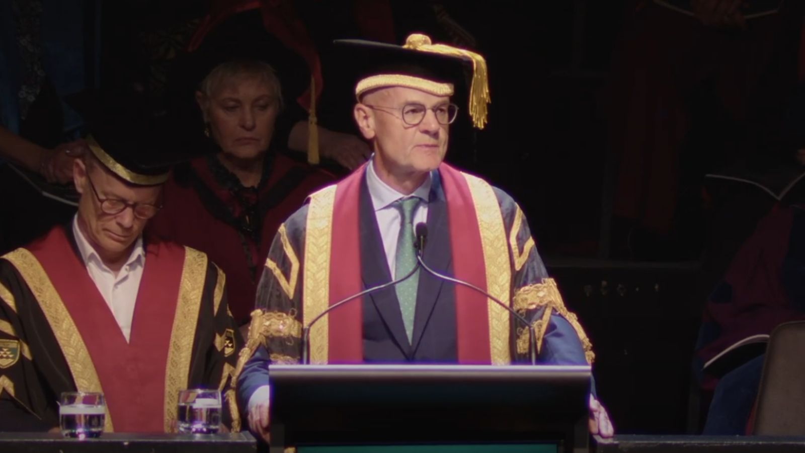 The Chancellor speaks at the graduation ceremony, with the Vice-Chancellor seated in the background.