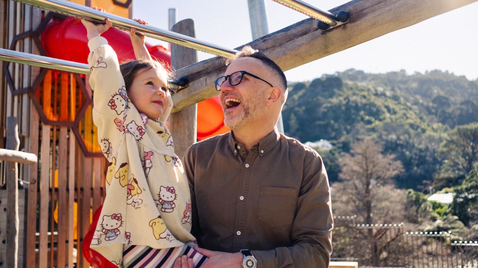 Mature student helping a young child across a set of monkey bars in a playground