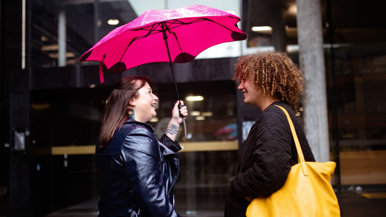 2 students at the entrance to a building in the rain, sharing an umbrella