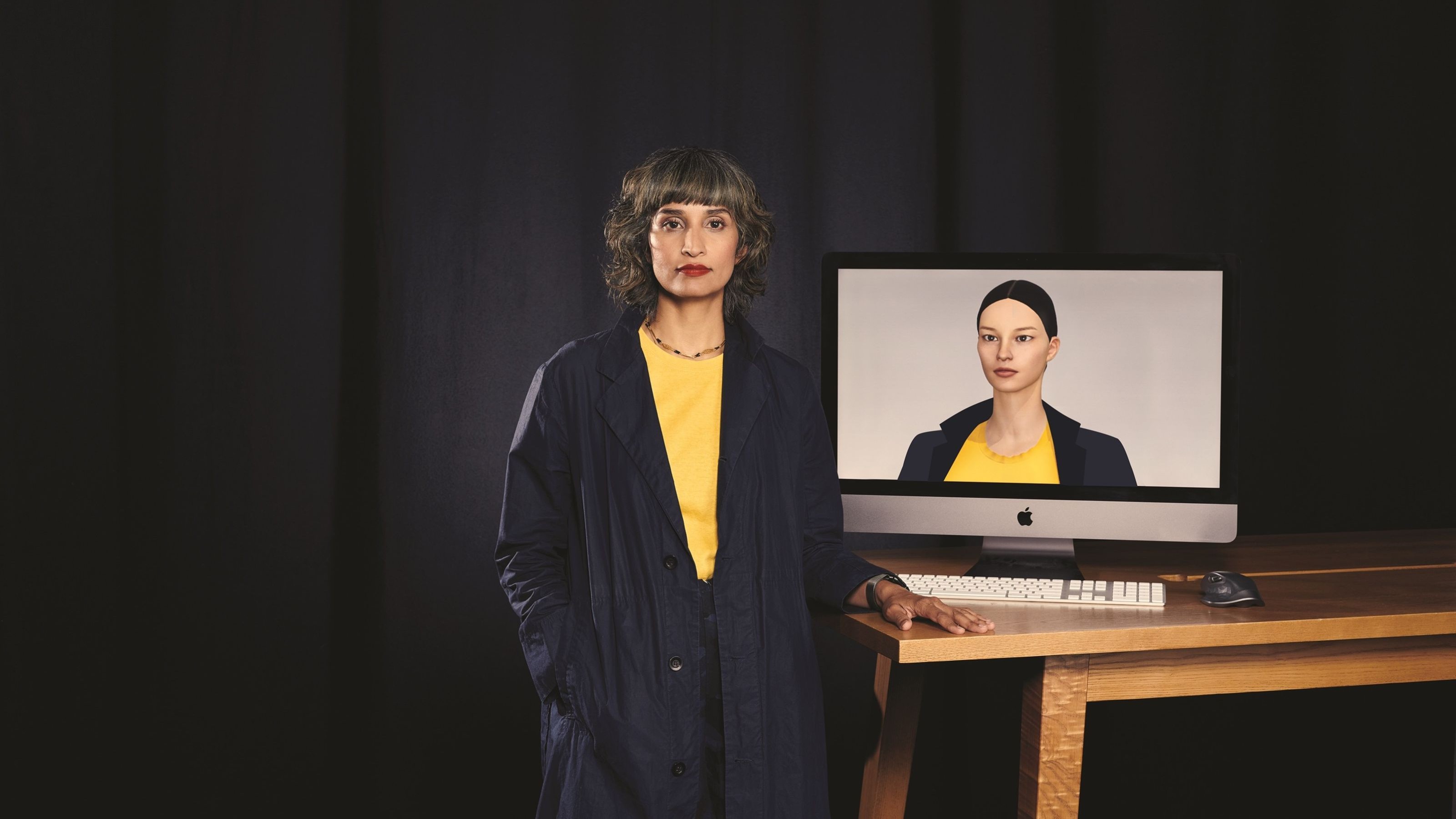 Raqi Syed standing in front of a computer screen displaying an animated woman with matching clothing. 