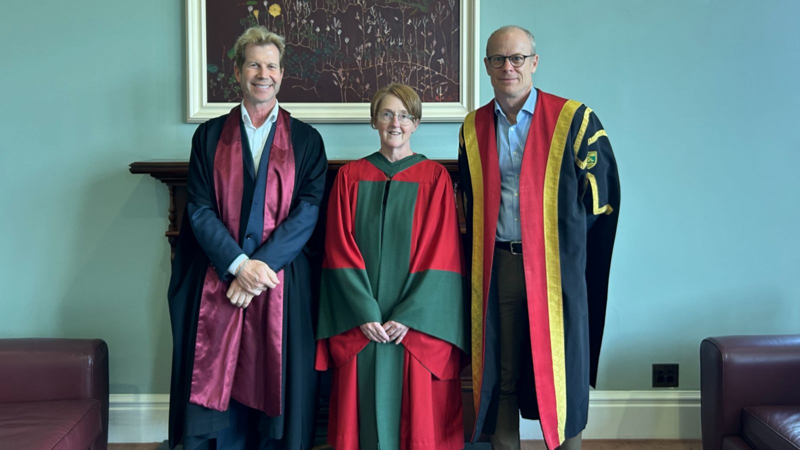 Three academics, two male, one female wearing academic dress in front of a pale green wall