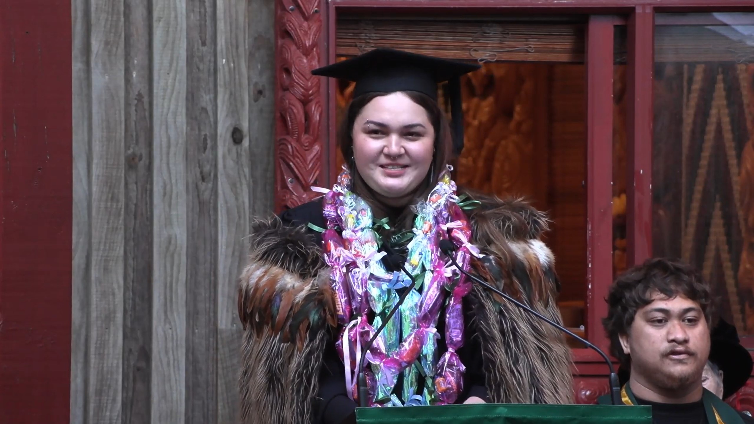 Graduate wearing academic gown and cap with colourful garlands, speaking at a podium.