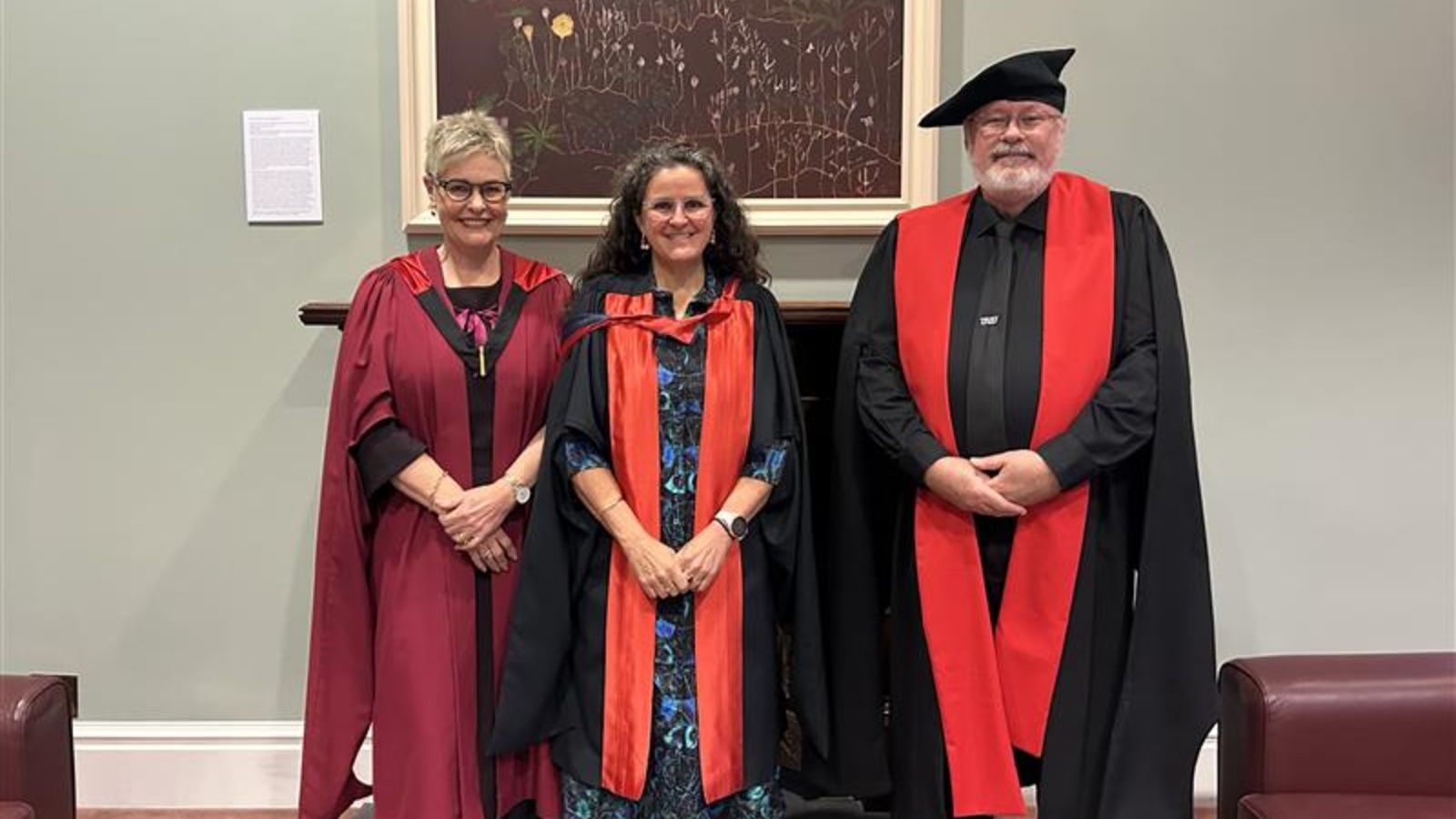 Three academics, two female, one male, wearing academic dress in front of a pale green wall