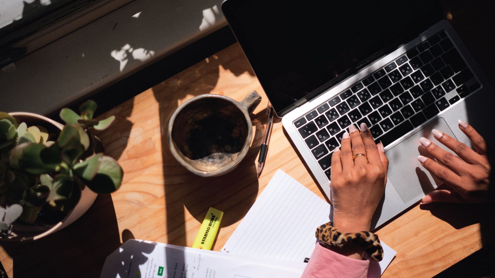 Top-down view of a sunny desk with a mug, plant, papers, and hands typing on a laptop.