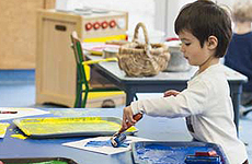Paint Time – A young boy in a children’s activity room, stands at a short desk with trays of yellow, green and blue paint. He holds a paint roller of blue paint and creates a masterpiece on white paper, all the while, looking quite pleased with his work.