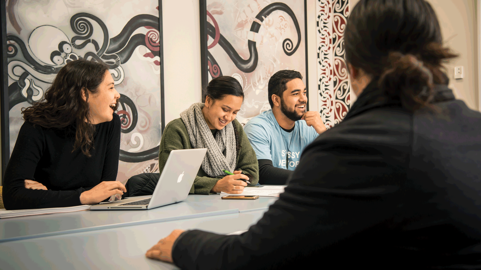 Students sit at a table with a laptop and enjoy a conversation – on the wall in the background, there is an artistic version of an octopus.