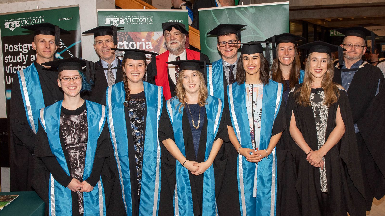 The eight meteorology graduates, wearing graduation gowns and caps, stand in front of Victoria banners with the MetService CEO and the Programme Director. Back row: Lewis Ferris, Peter Lennox (MetService CEO), James McGregor (Programme Director), Andrew James, Ashlee Parkes, Chris Webster (from MetService) Front row: Melissa Oosterwijk, Jessie Owen, Juliane Bergdolt, Tahlia Crabtree, Kathryn Boorman.