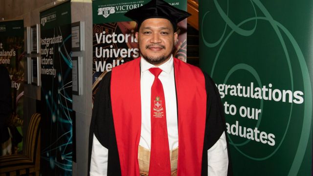PhD graduate Taitusi Taufa, in graduation attire, stands in front of Victoria University of Wellington graduation banners.