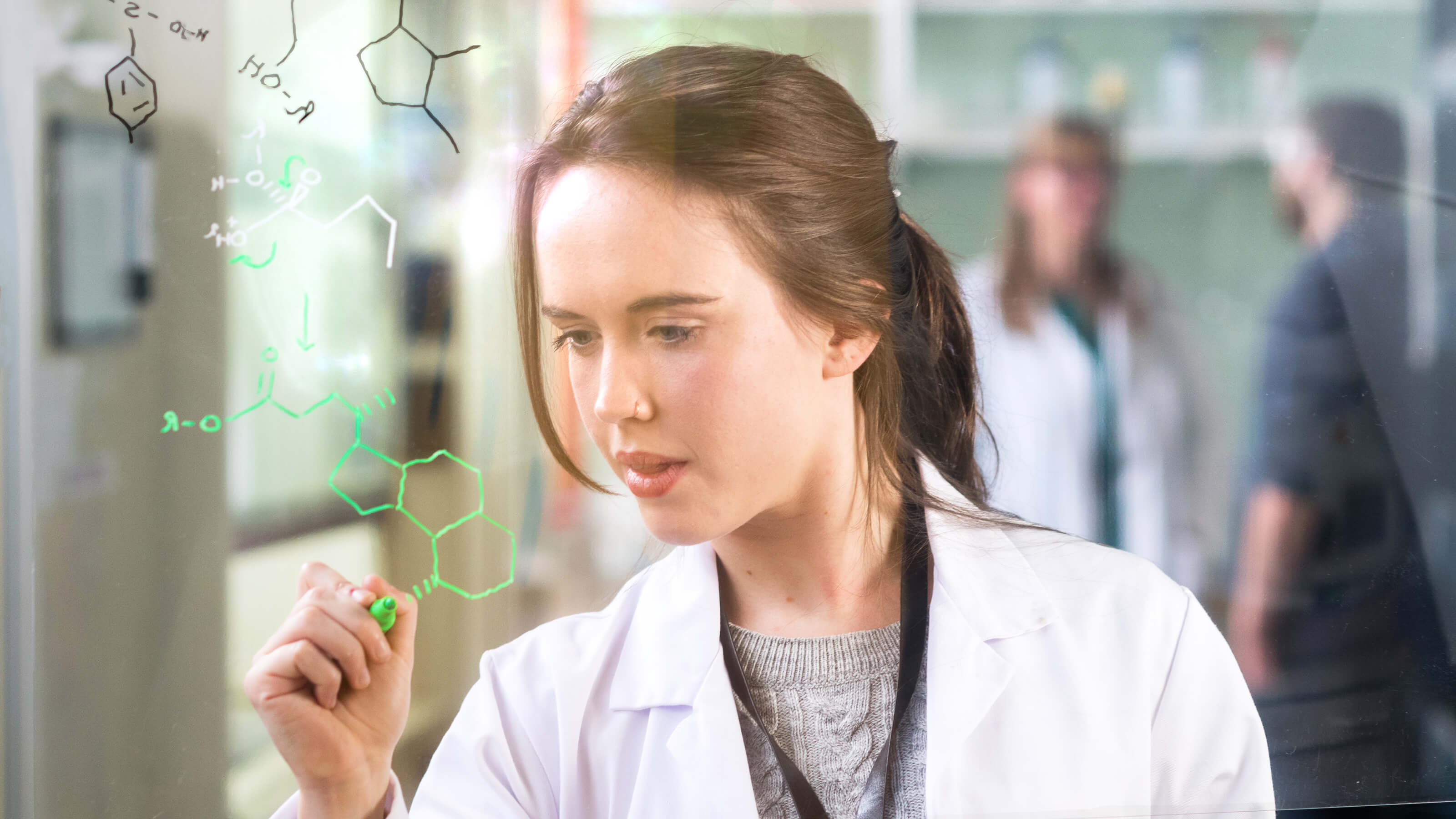 A female postgraduate science student is wearing a lab coat and writing formulas on a transparent whiteboard.