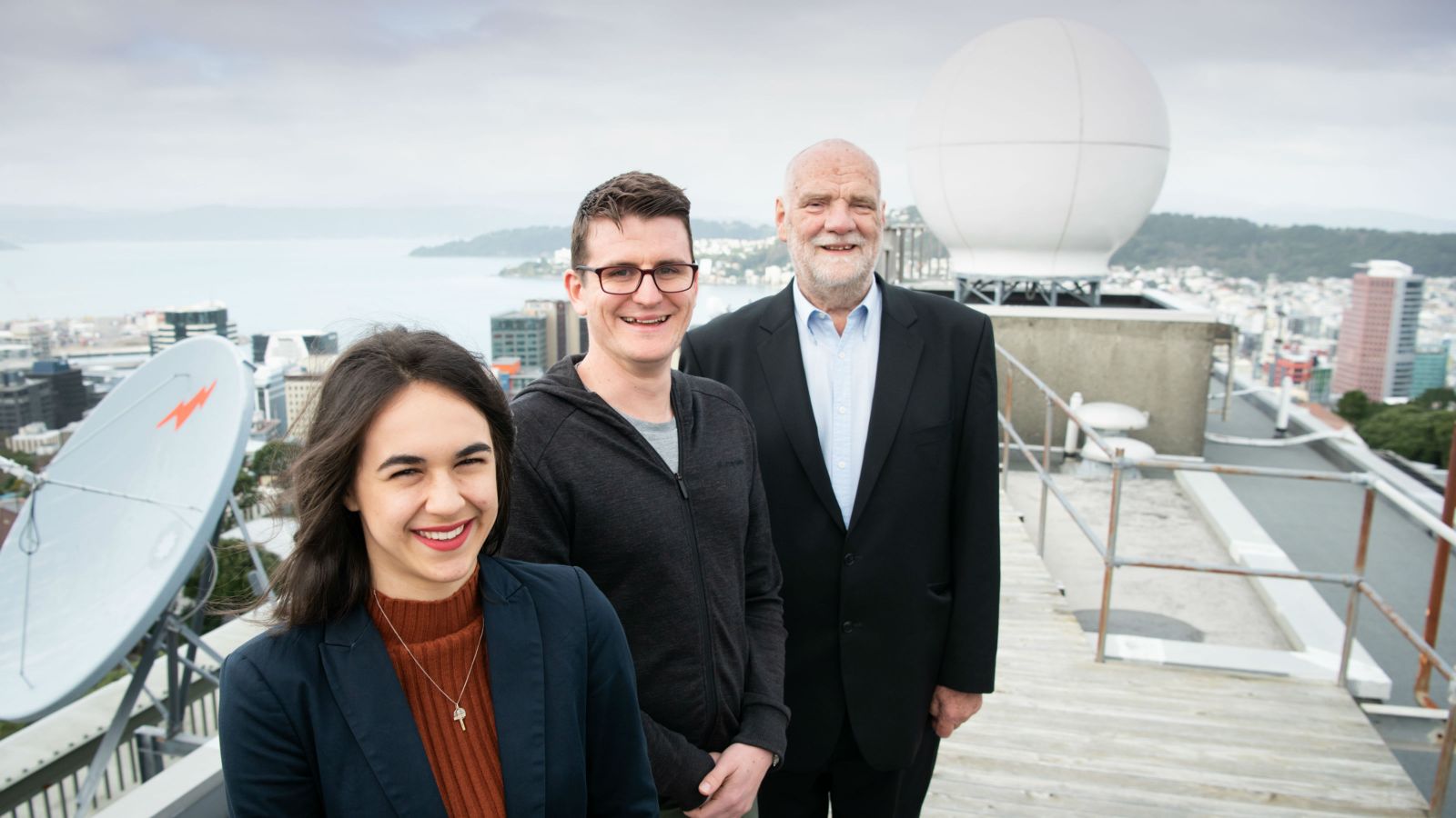 Tahlia Crabtree, Andrew James, and Dr Jim McGregor stand on the roof of the MetService, large satellite imagery equipment around them, and the coastline of Wellington harbour in the background.