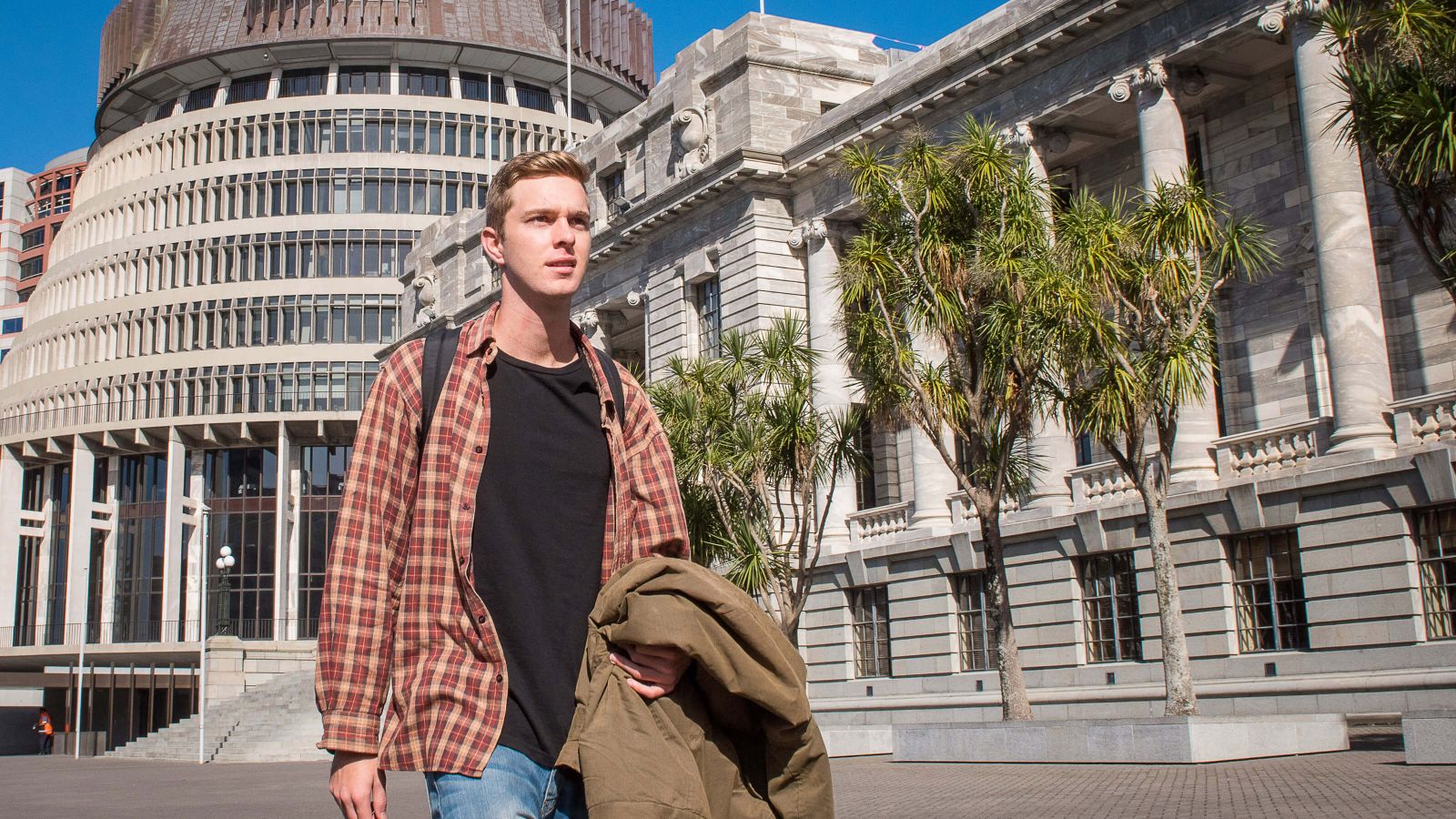 A young man dressed in a plaid shirt, black top, and blue jeans walks past the complex of New Zealand Parliament.   