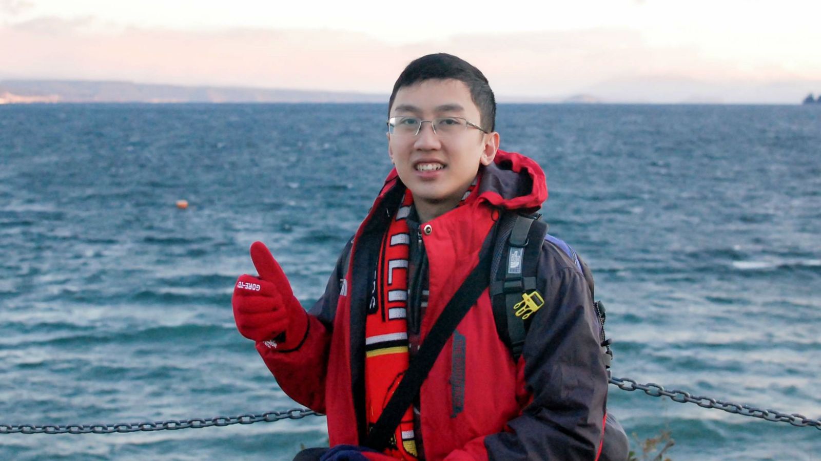 Quan Trong Nguyen in a big red jacket gives a gloved thumbs-up before a body of blue water and coastline in the offing.