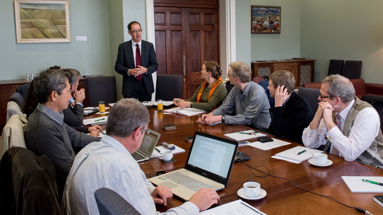 A man presents to a boardroom of people with laptops and paper in front of them.